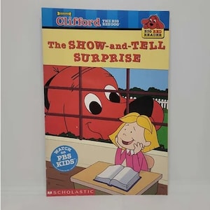 May include: A red dog named Clifford peeks through a window at a girl sitting at a desk with a book. The book is open and the girl is smiling. The book cover says "The Show-and-Tell Surprise". The book is part of the Clifford the Big Red Dog series.