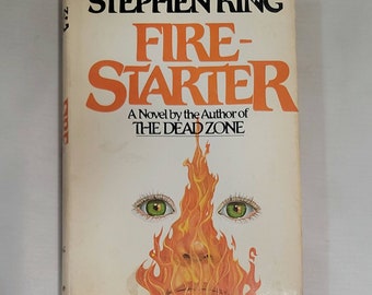May include: A paperback book cover with a white background and the title "Firestarter" in orange and black text. The author, Stephen King, is listed in black text above the title. The cover features an illustration of a face with green eyes and flames surrounding it.