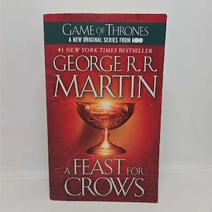 May include: A Feast for Crows, a novel by George R.R. Martin, is a #1 New York Times bestseller. The book cover features a golden goblet on a red background with the title in white text.