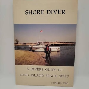 May include: A book titled "Shore Diver: A Divers Guide to Long Island Beach Sites" by Daniel Berg. The cover features a photo of a boat docked on a sandy shore with a diver standing nearby.