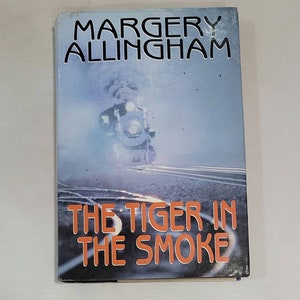 May include: A vintage paperback book cover with a blue background featuring a steam locomotive emerging from a cloud of smoke. The title "The Tiger in the Smoke" is printed in orange letters below the image. The author's name, "Margery Allingham", is printed in black letters at the top of the cover.