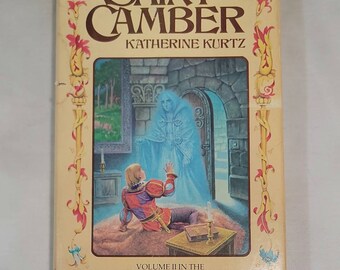 May include: A book cover with a yellow background and ornate gold trim. The title "Saint Camber" is in gold lettering with the author "Katherine Kurtz" below. The cover art features a man in a blue and red tunic looking up at a translucent figure in a white robe. The text at the bottom reads "Volume II in the Legends of Camber of Culdi."