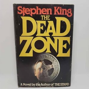 May include: A paperback book cover with a black background and yellow text. The title is "The Dead Zone" by Stephen King. The cover features a man's face with a clock face behind it. The text at the bottom reads "A Novel by the Author of THE STAND."