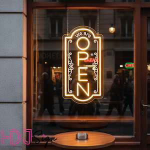Puede incluir: Un letrero de neón rectangular con bordes redondeados colgado en una ventana. El letrero muestra el texto "We are OPEN" en luz amarilla brillante, rodeado por un borde decorativo. Está suspendido por cadenas finas desde arriba.
