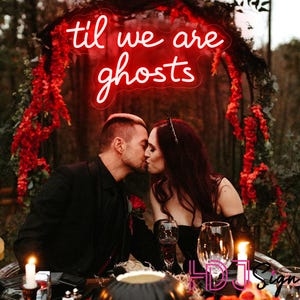 May include: A couple kissing at a candlelit table, with a red neon sign that reads "til we are ghosts." The scene is decorated with red flowers and a carved pumpkin. The overall theme is gothic and romantic.