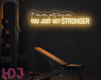 Op de afbeelding: Een aan de muur gemonteerd neonbord dat een warme gele gloed uitstraalt, met de tekst "It never gets easier. YOU JUST GET STRONGER" in een combinatie van cursieve en vetgedrukte schreefloze lettertypes, boven een zwarte gewichtsbank.