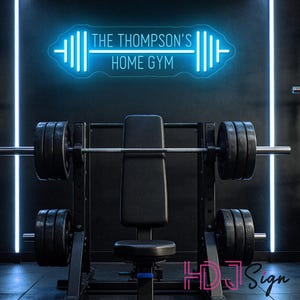 May include: A bright blue neon sign mounted on a dark wall featuring the text THE THOMPSON'S HOME GYM centred within a stylized barbell graphic. The sign emits a cool glow above a black weight bench and barbell rack setup in a gym environment.