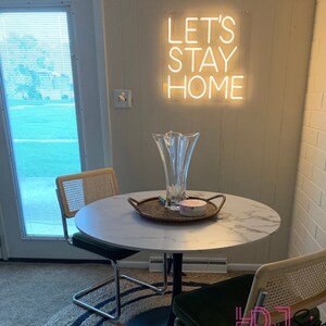 May include: A white marble table with a black metal base, two chairs with woven cane backs, and a glass vase with a flower arrangement. The table is decorated with a woven tray and a small pink box. A neon sign on the wall reads "Let's Stay Home".