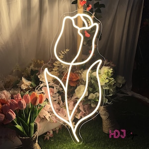 May include: White neon sign shaped like a tulip flower with a stem and leaves. The sign is lit up and is in front of a background of flowers.