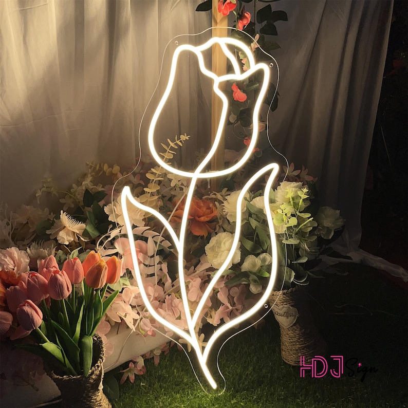 May include: White neon sign shaped like a tulip flower with a stem and leaves. The sign is lit up and is in front of a background of flowers.