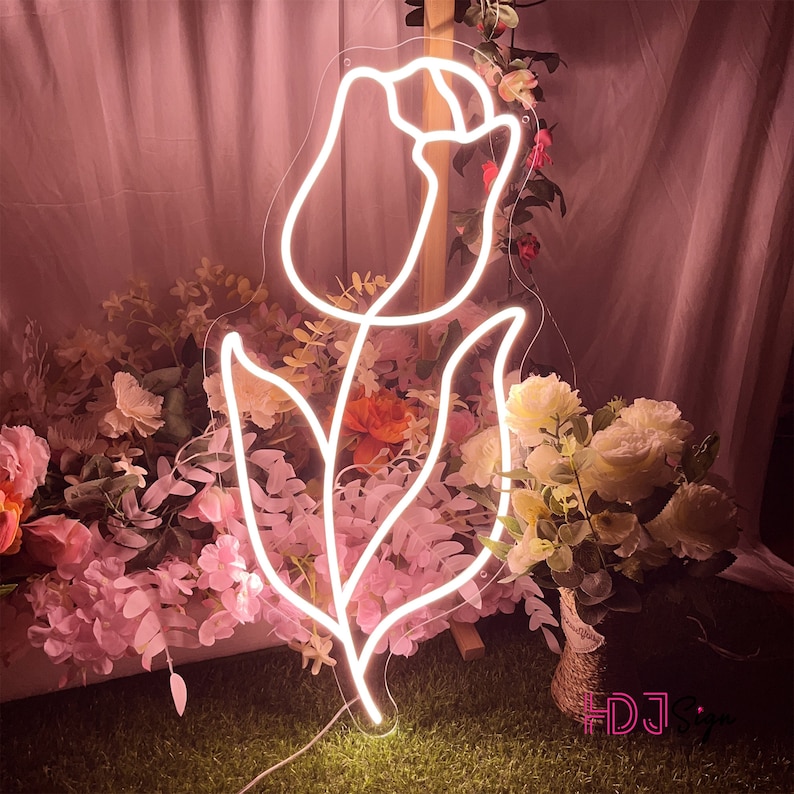 May include: A white neon sign shaped like a tulip flower with a stem and leaves. The sign is lit up and is sitting on a table with pink and white flowers.