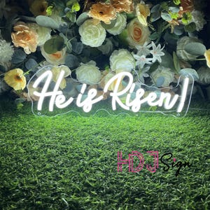 May include: White neon sign with the text "He is Risen!" in a cursive font. The sign is on a green grass background with white flowers.