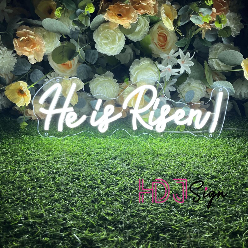 May include: White neon sign with the text "He is Risen!" in a cursive font. The sign is on a green grass background with white flowers.