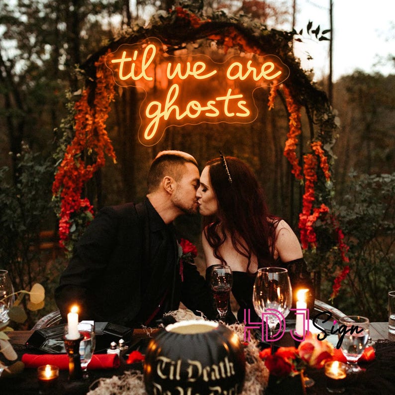 May include: A neon sign with the words "til we are ghosts" in a cursive font, above a couple kissing. The scene is set outdoors with a decorated archway and a table with candles and a carved pumpkin.