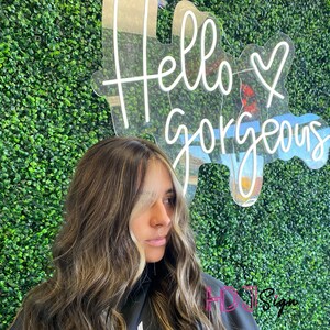 May include: A woman with long, wavy, light brown hair with blonde highlights stands in front of a green wall with a neon sign that reads "Hello Gorgeous".