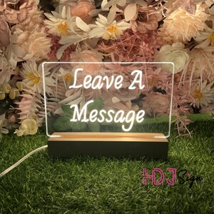 Op de afbeelding: Een wit acryl bord met een houten basis, verlicht van achteren. Het bord heeft de tekst "Leave A Message" in een cursief schrift.