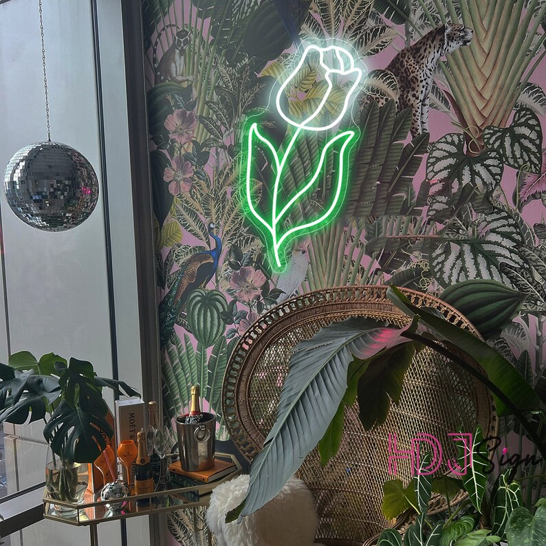 May include: A neon green floral sign in the shape of a tulip with a white outline hangs on a wall with a tropical print wallpaper. The sign is illuminated and casts a soft glow.