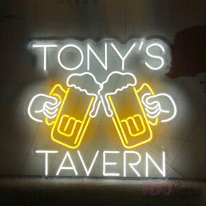 Op de afbeelding: Wit neon-bord met de tekst "TONY'S TAVERN" en twee gele bierglazen met wit schuim, die tegen elkaar klinken.