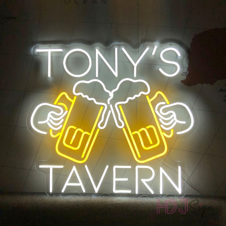 Op de afbeelding: Wit neon-bord met de tekst "TONY'S TAVERN" en twee gele bierglazen met wit schuim, die tegen elkaar klinken.
