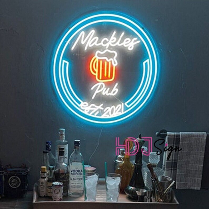 May include: A blue neon sign with white lettering that reads "Mackles Pub est 2021". The sign features a stylised beer mug with orange and yellow accents. The sign is mounted on a wall above a bar counter with bottles and glasses.