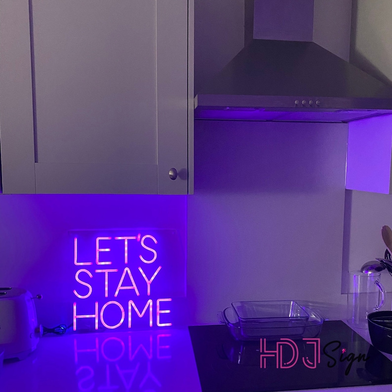 May include: Pink neon sign with the text "Let's Stay Home" on a white background with purple lighting. The sign is reflected in a glass surface.