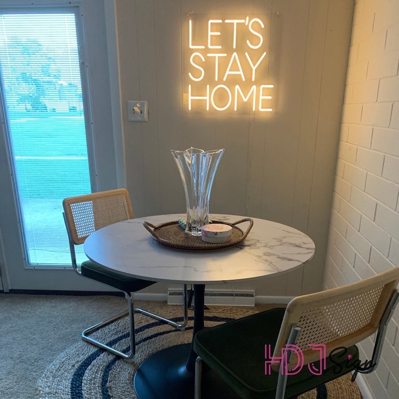 May include: A white marble table with a black metal base, two chairs with woven cane backs, and a glass vase with a flower arrangement. The table is decorated with a woven tray and a small pink box. A neon sign on the wall reads "Let's Stay Home".
