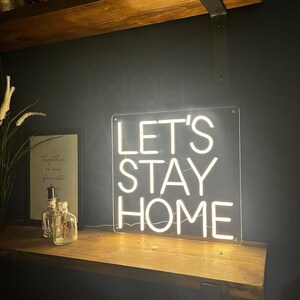 May include: White neon sign with the words "Let's Stay Home" on a black background. The sign is mounted on a wooden shelf.