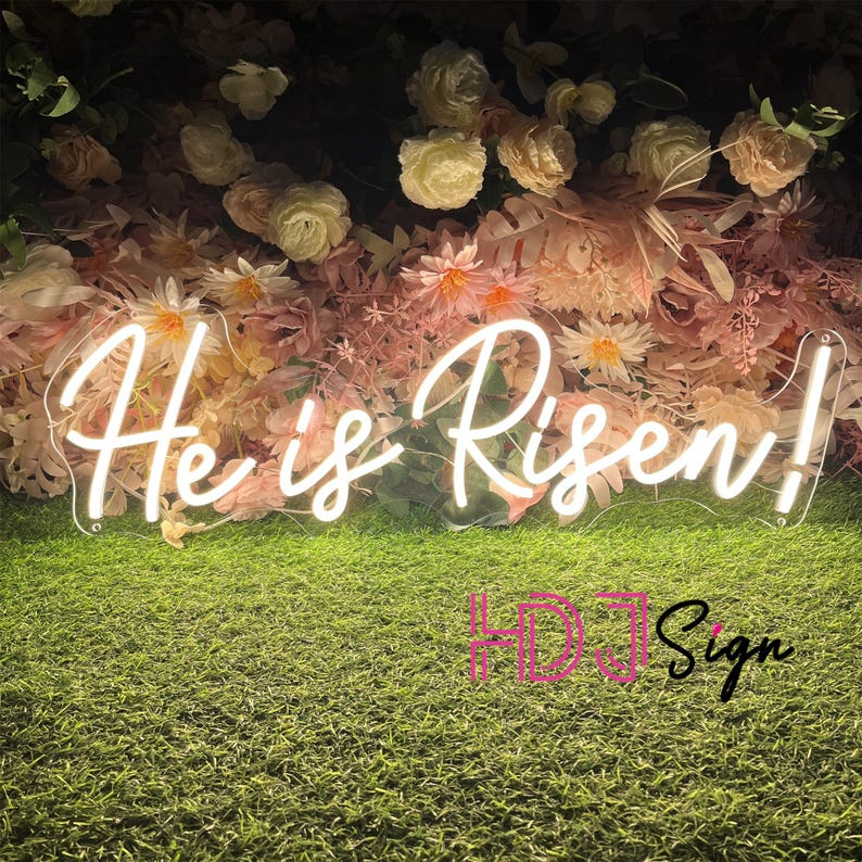 May include: White neon sign with the text "He is Risen!" in a cursive font. The sign is in front of a wall of pink and white flowers.