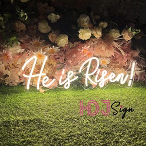 May include: White neon sign with the words "He is Risen!" against a backdrop of pink and white flowers.