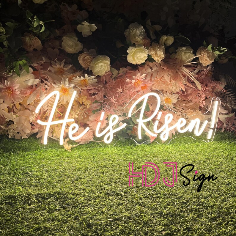 May include: White neon sign with the words "He is Risen!" against a backdrop of pink and white flowers.