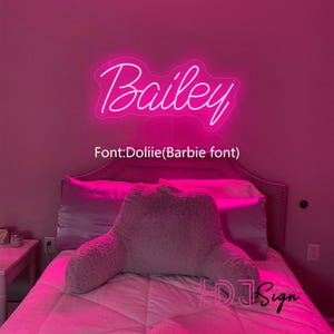 Op de afbeelding: Een roze slaapkamer met een wit neon-bord met daarop "Bailey" in een cursief schrift. De tekst "Font: Dollie (Barbie font)" staat onder het bord. Het bed heeft een wit kussen en een roze kussen.