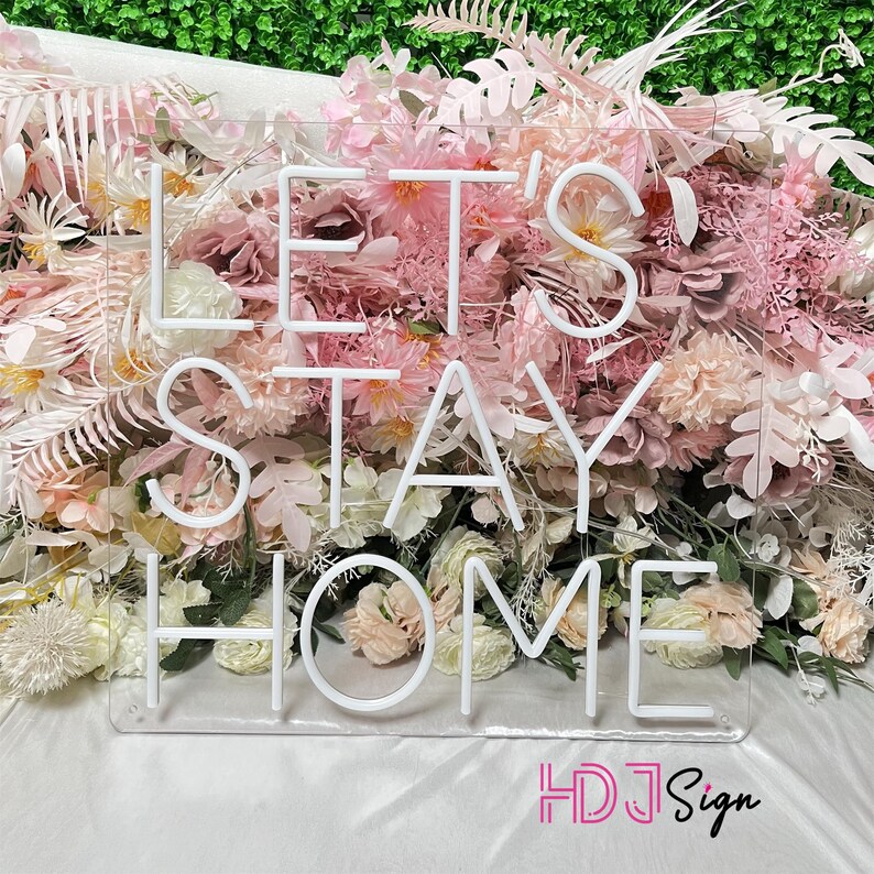May include: White neon sign with the words "Let's Stay Home" on a clear acrylic background. The sign is surrounded by pink and white flowers.