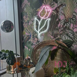 May include: A pink neon light shaped like a rose with a green and pink floral wallpaper background. A wicker chair with a green plant in front of it.