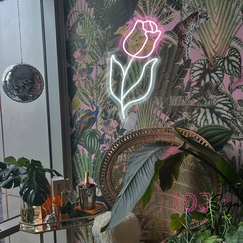 May include: A pink neon light shaped like a rose with a green and pink floral wallpaper background. A wicker chair with a green plant in front of it.
