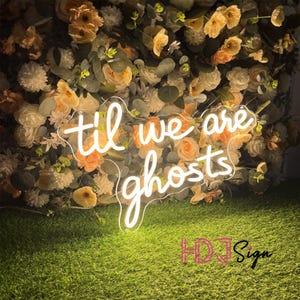 May include: A neon sign with the words "til we are ghosts" in a cursive font. The sign is white and sits in front of a wall of flowers in shades of peach, cream, and green. The sign is on a bed of green grass.