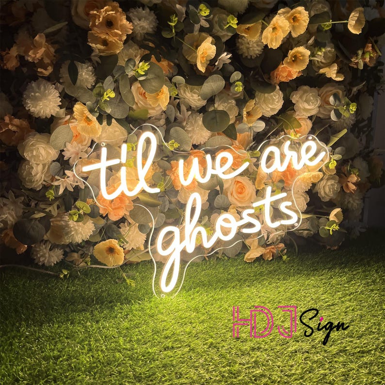 May include: A neon sign with the words "til we are ghosts" in a cursive font. The sign is white and sits in front of a wall of flowers in shades of peach, cream, and green. The sign is on a bed of green grass.