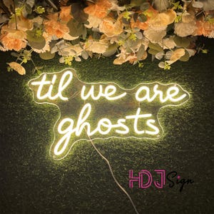 May include: A neon sign with the words "til we are ghosts" in a cursive font, illuminated in a soft white glow. The sign is set against a backdrop of artificial green grass and adorned with peach and white flowers.