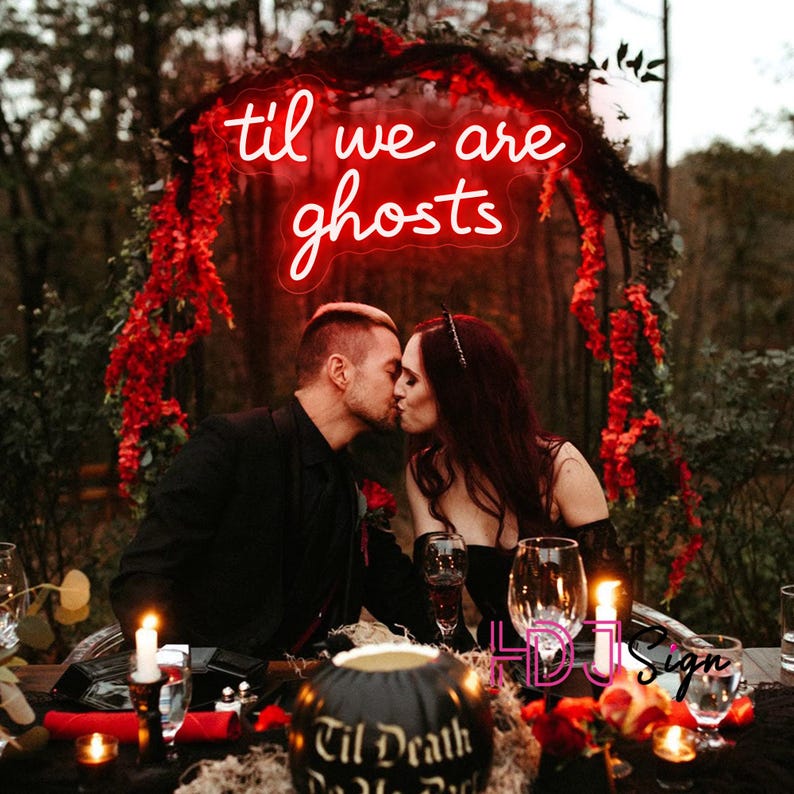 May include: A couple kissing at a candlelit table, with a red neon sign that reads "til we are ghosts." The scene is decorated with red flowers and a carved pumpkin. The overall theme is gothic and romantic.