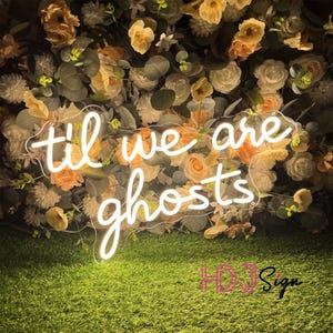 May include: A neon sign with the words "til we are ghosts" in a cursive font. The sign is white and set against a backdrop of cream and peach-colored flowers and greenery. The sign is placed on a bed of green artificial grass.