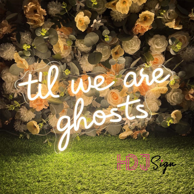 May include: A neon sign with the words "til we are ghosts" in a cursive font. The sign is white and set against a backdrop of cream and peach-colored flowers and greenery. The sign is placed on a bed of green artificial grass.
