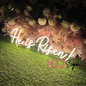 May include: White neon sign with the words "He is Risen!" in a cursive font. The sign is placed in front of a wall of pink and white flowers.