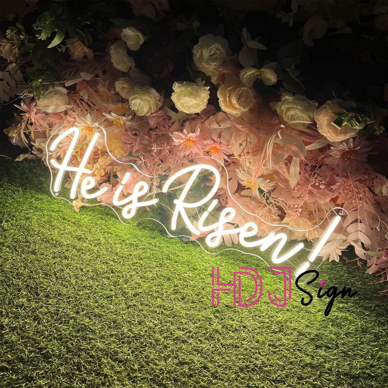 May include: White neon sign with the words "He is Risen!" in a cursive font. The sign is placed in front of a wall of pink and white flowers.