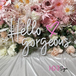May include: White neon sign with the text "Hello gorgeous" and a pink neon heart. The sign is displayed against a backdrop of pink and white flowers.