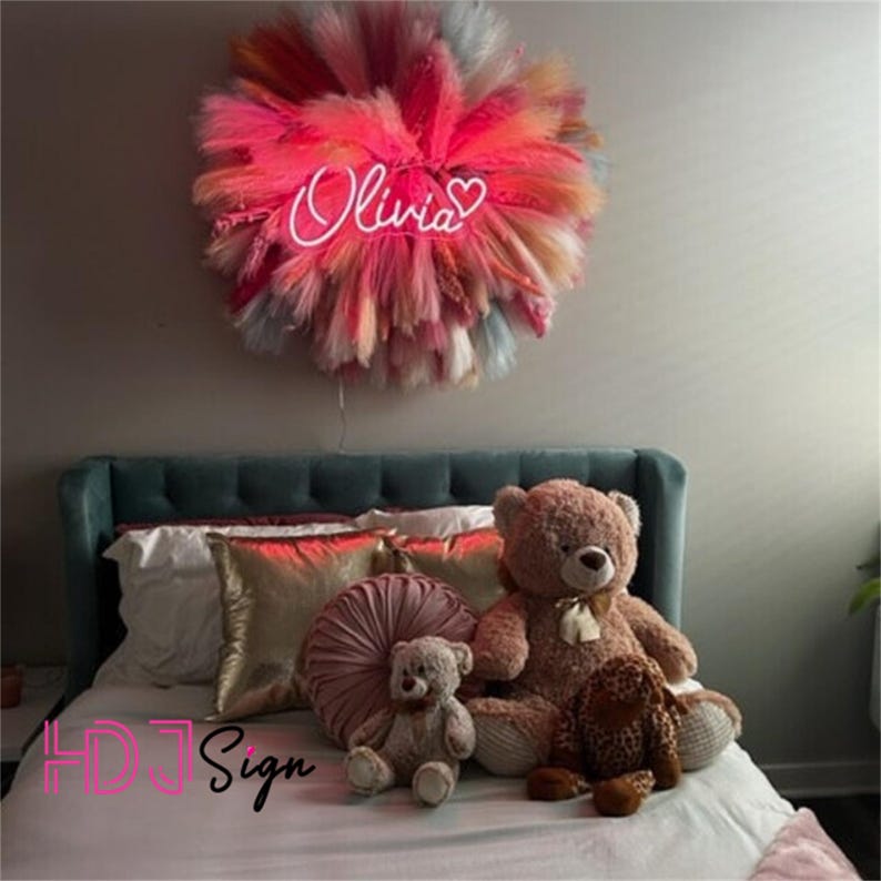 Op de afbeelding: Een teal hoofdbord met een wit beddengoed en roze kussens. Een grote teddybeer zit op het bed met een kleinere teddybeer ervoor. Een roze neonbord met de naam "Olivia" en een hart hangt aan de muur boven het bed.