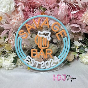 May include: A neon sign with a blue and white circular border. The sign says "Savage Bar Est 2024" in white neon lettering. Inside the circle is a neon orange beer mug with a white foam top.