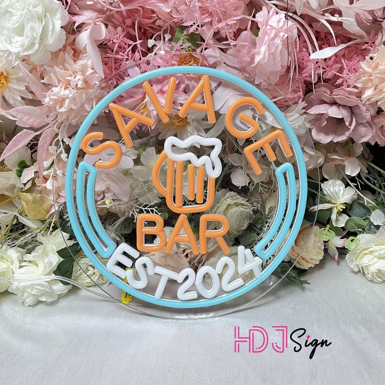 May include: A neon sign with a blue and white circular border. The sign says "Savage Bar Est 2024" in white neon lettering. Inside the circle is a neon orange beer mug with a white foam top.