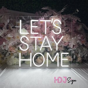 May include: White neon sign with the text "Let's Stay Home" on a clear acrylic background. The sign is surrounded by pink flowers.