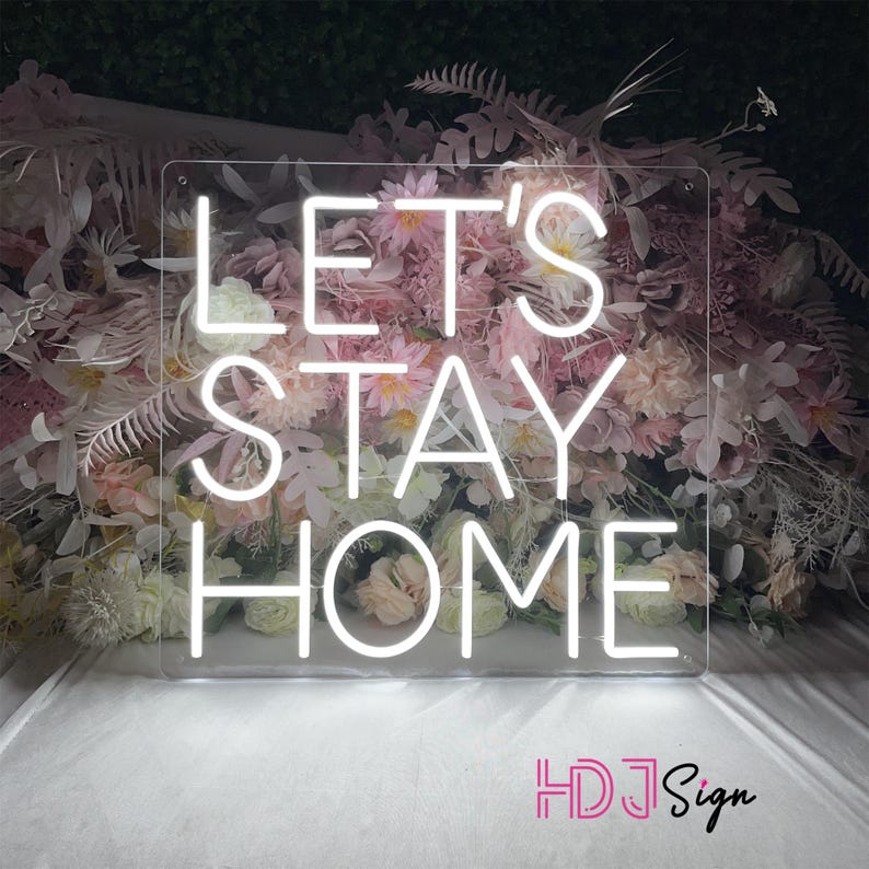 May include: White neon sign with the text "Let's Stay Home" on a clear acrylic background. The sign is surrounded by pink flowers.