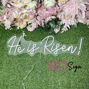 May include: White neon sign with the words "He is Risen!" on a green grass background. The sign is made of clear acrylic and has a white neon light.