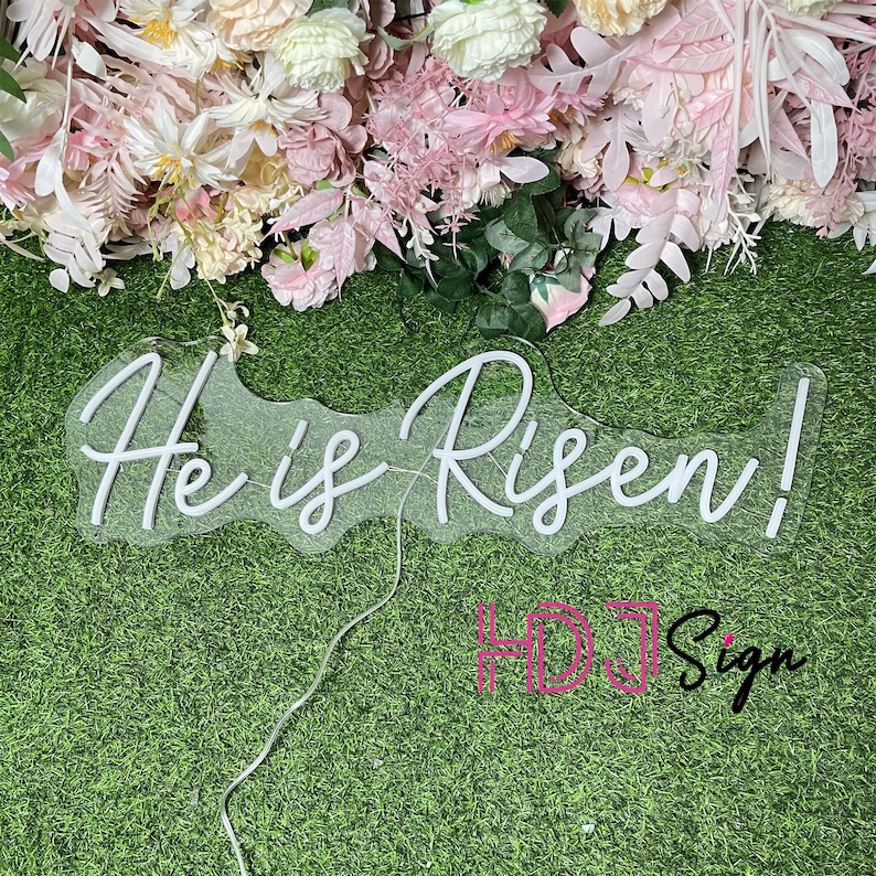 May include: White neon sign with the words "He is Risen!" on a green grass background. The sign is made of clear acrylic and has a white neon light.